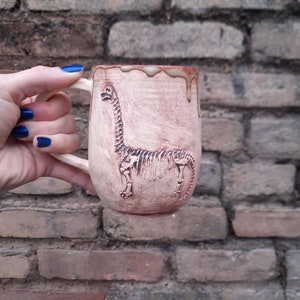 Könnte beinhalten: Eine handgefertigte Keramik-Tasse mit einem Dinosaurier-Skelett-Design. Die Tasse ist hellbraun mit einer strukturierten Oberfläche und einem Henkel. Das Dinosaurier-Skelett ist in die Seite der Tasse geschnitzt.