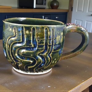 Puede incluir: Taza de cerámica hecha a mano con un diseño único. La taza presenta un esmalte verde oscuro y azul con un patrón texturizado y tallado. El asa tiene un detalle decorativo texturizado. Una pieza funcional y artística.