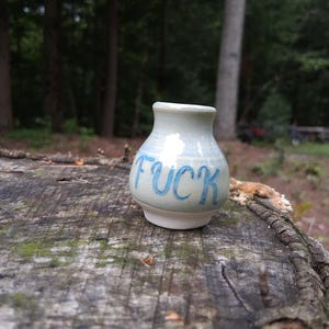 May include: A small, light blue ceramic vase with a glossy finish and a narrow neck. The word FUCK is hand-painted in blue lettering across the front of the vase. The base of the vase is unglazed, showing a matte white ceramic texture.