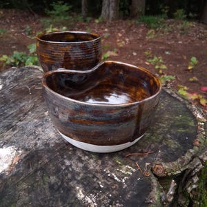 May include: Two handmade ceramic bowls with a glossy finish. The front bowl features a heart-shaped rim, while the back bowl is cylindrical. Both pieces display a blend of deep brown, amber, and grey tones with a solid white base at the bottom.