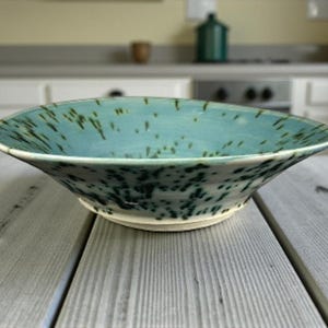 Puede incluir: Un cuenco de cerámica hecho a mano con un diseño ancho y poco profundo. El cuenco presenta un esmalte turquesa con motas verde oscuro y una base blanca. Se encuentra sobre una mesa de madera gris, mostrando su estilo artístico único.