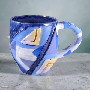 Puede incluir: Una taza de cerámica con interior y exterior azules, con un diseño geométrico en tonos azules, blancos y amarillos. El asa también está decorada con patrones azules y blancos. La taza tiene una capacidad de aproximadamente 350 ml.