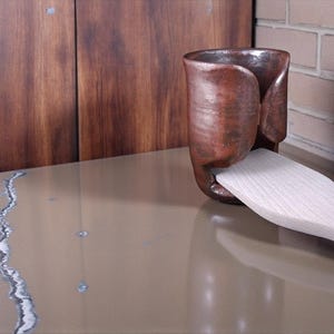 Puede incluir: Un soporte para utensilios de cerámica de color cobre hecho a mano con una espátula de madera. El soporte tiene un diseño curvo único y se coloca sobre una superficie de tonos neutros. La espátula es de madera clara.