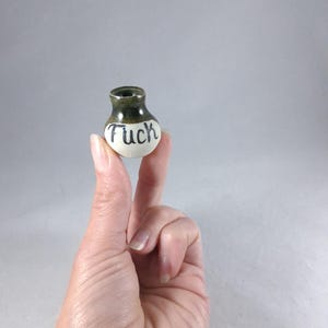 May include: A small, round ceramic vase with a narrow neck and a wide base. The upper portion is glazed in a dark olive green, while the lower portion is off-white with the word "Fuck" written in black, cursive, hand-painted lettering on the front.