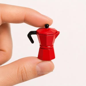 Può includere: Una piccola caffettiera rossa con manico e parte superiore neri. Il design geometrico in miniatura è tenuto tra due dita. La caffettiera è di un colore rosso vibrante e ha un classico design italiano.