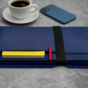 Può includere: Una custodia blu navy per laptop con una fascia elastica nera, contenente un blocco note giallo e una penna rossa. Una tazza da caffè bianca e uno smartphone blu sono sulla superficie grigia. La custodia è progettata per proteggere un laptop.