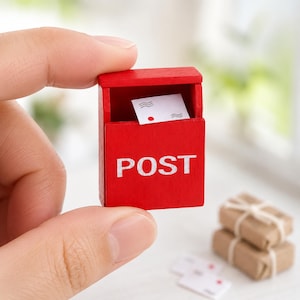 Pode incluir: Uma caixa de correio em miniatura de madeira vermelha brilhante com a palavra POST impressa em branco na frente. A caixa de correio tem uma ranhura superior aberta contendo um pequeno envelope branco com linhas onduladas pretas e um ponto.