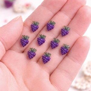Puede incluir: Diez dijes en miniatura de uvas moradas con hojas verdes. Los dijes son pequeños y detallados, posiblemente de resina, y están dispuestos sobre una superficie rosa claro.