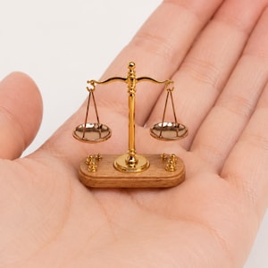 Peut inclure: Petite balance dorée avec deux plateaux circulaires et un support central. La balance repose sur une petite base en bois avec des détails dorés. La balance est tenue dans la paume ouverte d'une personne.