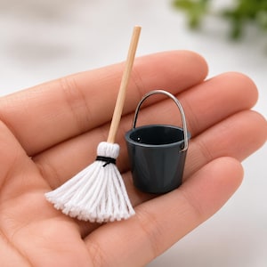 May include: A miniature cleaning set consisting of a small mop with a light wooden handle and white cotton strands tied with a black thread, alongside a tiny dark gray bucket featuring a silver-toned metal handle attached to the sides.