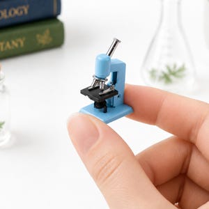 Può includere: Un microscopio in miniatura azzurro con dettagli argentati e neri è tenuto in mano. Il piccolo microscopio ha una base, un tavolino e un oculare. Lo sfondo include libri e un'ampolla di vetro, suggerendo un tema scientifico o educativo.