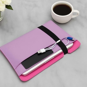 May include: A lavender tablet sleeve with a black elastic band closure, holding a tablet, a black phone, and a white charging cable. The sleeve has a pink interior. A cup of coffee and a vase of white flowers are in the background.