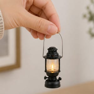 May include: A miniature black lantern with a clear glass enclosure and a silver handle. The lantern is lit with a warm, yellow light, suggesting a cozy, vintage aesthetic. The lantern is held in a hand.