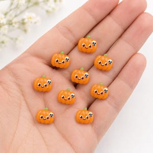 Könnte beinhalten: Eine Ansammlung von neun kleinen, orangefarbenen kürbisförmigen Anhängern. Jeder Kürbis zeigt ein lächelndes Gesicht mit großen Augen und ein kleines weißes Totenkopf-Symbol an der Seite. Die Anhänger haben eine glatte, glänzende Oberfläche.
