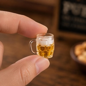 Pode incluir: Um pequeno copo de cerveja de vidro cheio de líquido dourado e coberto com espuma branca. O pequeno copo, com uma alça, é segurado entre dois dedos. O fundo está desfocado, sugerindo um pub ou bar.
