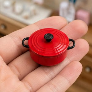 May include: A miniature red cooking pot with a black handle and a black knob on the lid. The small pot is round and has a glossy finish. The pot is a kitchen-themed item.