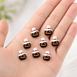 May include: Eight small, black, glossy pumpkin-shaped charms, each featuring a carved orange jack-o'-lantern face and a white top hat with a thin red band. These miniature decorative pieces are arranged in a cluster on a flat surface.
