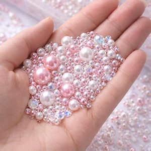 May include: A collection of pink and white pearls and iridescent beads are held in a hand. The pearls vary in size, with larger pink and white pearls interspersed among smaller ones. The beads are scattered around the hand.