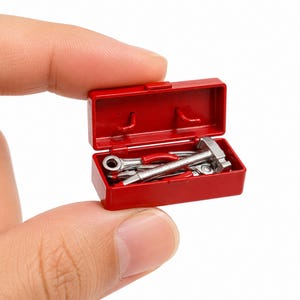Pode incluir: Uma caixa de ferramentas em miniatura de plástico vermelho com a tampa aberta, contendo uma pequena coleção de ferramentas manuais de metal prateado, incluindo um martelo e chaves, apresentada num estojo retangular compacto.