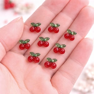 Puede incluir: Pequeñas cuentas rojas en forma de cereza con hojas verdes sobre una mano. Las cuentas son brillantes y parecen ser de vidrio. Las cerezas son de un color rojo vibrante.