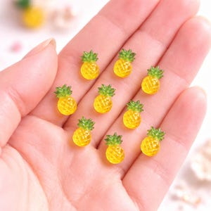 May include: A collection of small, yellow pineapple charms with green leafy tops. The miniature pineapples are arranged on a light background, showcasing their detailed design. These charms are ideal for crafting or jewelry making.