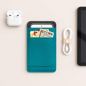 Puede incluir: Funda de teléfono de cuero color turquesa con una parte superior de fieltro gris, que contiene un teléfono inteligente, tarjetas de crédito y un billete de cincuenta dólares. También se ve un cable de carga blanco y auriculares inalámbricos.