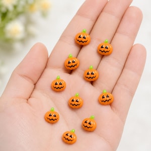 May include: A collection of ten small, orange, pumpkin-shaped charms, each featuring a black painted jack-o'-lantern face and a tiny green stem on top. These miniature decorative pieces are smooth, glossy, and uniform in their design.