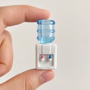 May include: A miniature white water cooler with a blue water jug on top. The cooler has pink and blue spouts. The item is held between fingers.