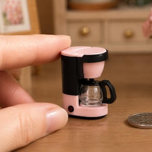 May include: A miniature pink and black coffee maker with a glass carafe and black handle. The small appliance has a red power button. A coin is in the foreground, indicating the scale of the item. The coffee maker is held by a hand.