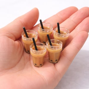 Könnte beinhalten: Miniatur-Bubble-Tea-Getränke mit brauner Flüssigkeit, schwarzen Strohhalmen und dunklen Tapiokaperlen. Die kleinen, durchsichtigen Becher haben einen hellbraunen Rand. Die Getränke werden in einer Hand vor weißem Hintergrund gehalten.