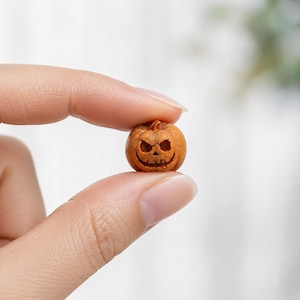 Può includere: Una miniatura di zucca arancione con un volto intagliato da jack-o'-lantern, occhi triangolari e un ampio sorriso sdentato. Il piccolo oggetto rotondo presenta una superficie ruvida e un picciolo sulla parte superiore.