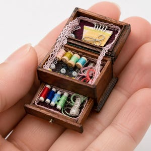Puede incluir: Caja de costura de madera en miniatura con dos cajones, llena de carretes de hilo de colores, agujas, tijeras y una cinta métrica. La caja está adornada con encaje y contiene cuentas y otros accesorios de costura. La caja se sostiene en una mano.