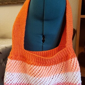 May include: A crocheted tote bag with a rainbow color pattern. The bag is made with orange, pink, white, and red yarn. The bag has a wide strap and is large enough to carry everyday items.