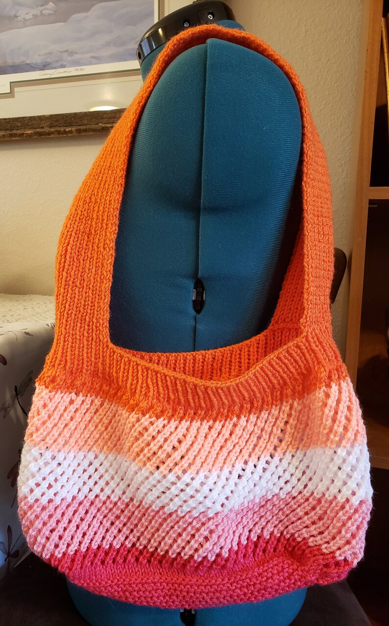 May include: A crocheted tote bag with a rainbow color pattern. The bag is made with orange, pink, white, and red yarn. The bag has a wide strap and is large enough to carry everyday items.