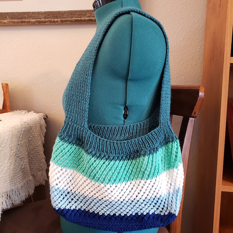 May include: A knitted tote bag with a teal, green, white, blue, and navy blue striped pattern. The bag has a wide, flat bottom and a single strap.