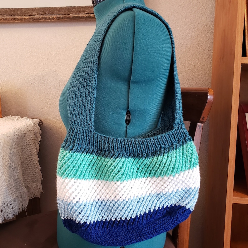 May include: A knitted tote bag with a teal, green, white, blue, and navy blue striped design. The bag has a wide, flat base and a single strap.