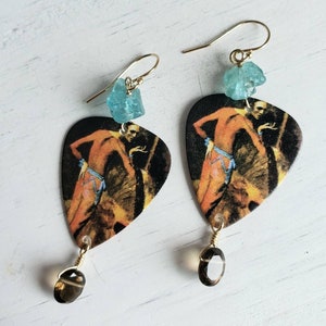 Op de afbeelding: Een paar oorbellen met gitaarplectrums met een vintage afbeelding van een dansende vrouw. De plectrums zijn versierd met een lichtblauwe edelsteen en een bruine edelsteen.