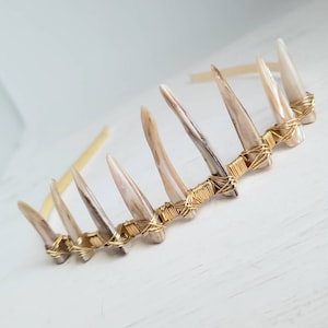 Puede incluir: Una diadema de alambre dorado con una fila de conchas blancas iridiscentes unidas a ella. Las conchas están dispuestas en un patrón escalonado y están unidas con alambre dorado.