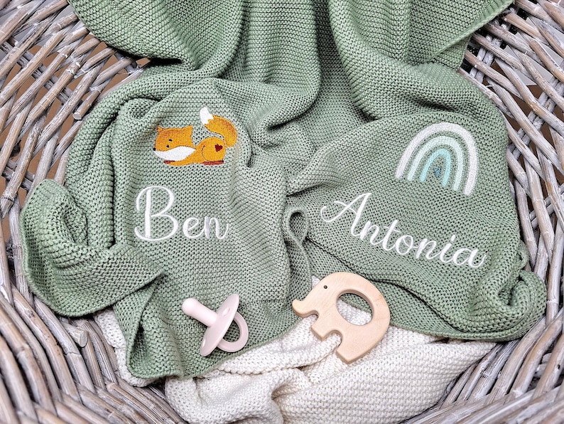Salbeigrüne Babydecken aus Strick mit Namen bestickt. Eine Decke zeigt einen Fuchs, die andere einen Regenbogen. Die Namen "Ben" und "Antonia" sind weiß gestickt.