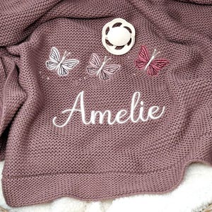 May include: A mauve knitted baby blanket featuring three embroidered butterflies in white, light pink, and dark pink. The name Amelie is embroidered in white cursive script below the butterflies, and a white pacifier rests on the fabric.