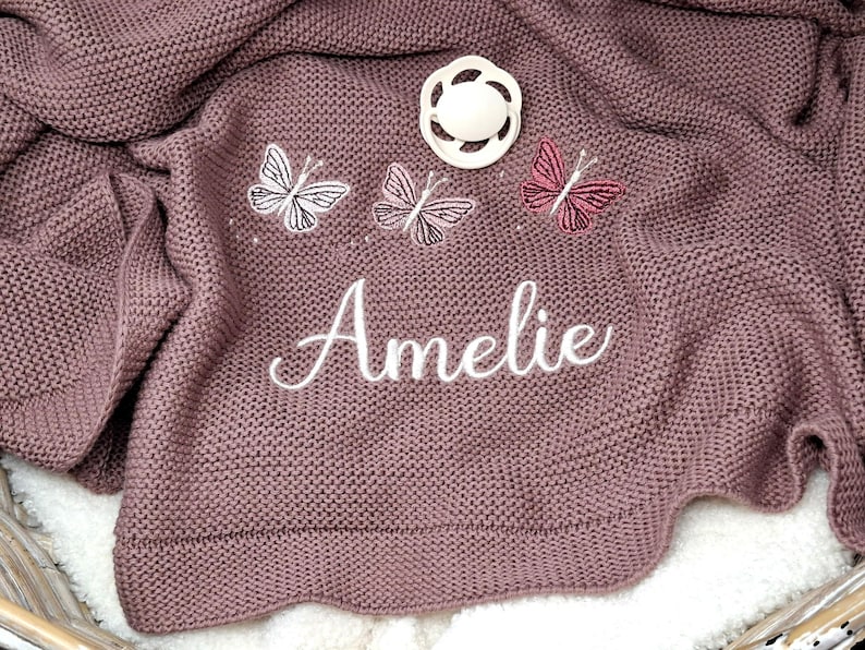May include: A mauve knitted baby blanket featuring three embroidered butterflies in white, light pink, and dark pink. The name Amelie is embroidered in white cursive script below the butterflies, and a white pacifier rests on the fabric.