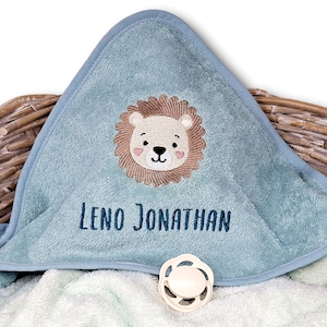 May include: A light blue hooded baby towel with an embroidered lion face and the name "Leno Jonathan" in navy blue. A white pacifier is also visible. The towel is folded over a wicker basket.