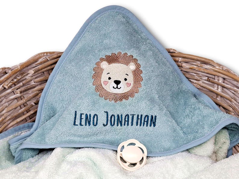 May include: A light blue hooded baby towel with an embroidered lion face and the name "Leno Jonathan" in navy blue. A white pacifier is also visible. The towel is folded over a wicker basket.