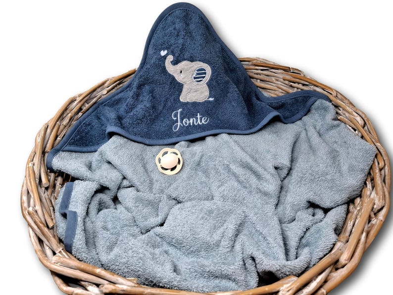 May include: A navy blue hooded baby towel with an embroidered elephant design and the name "Jonte" in white script. The towel is folded in a wicker basket with a gray towel underneath. A wooden pacifier is also in the basket.
