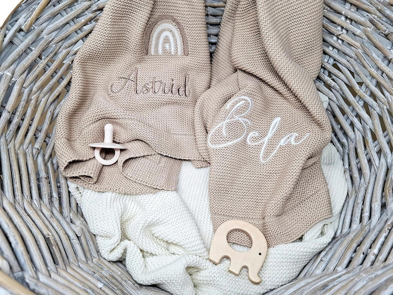 Gestrickte Babydecken mit Namen und Motiv bestickt. Eine mit "Astrid" und einem Regenbogen bestickt, die andere Babydecke ist mit dem Namen "Bela" bestickt.