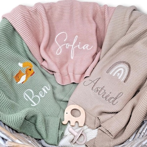 Drei gestrickte Babydecken mit Name und Motiv bestickt. Eine ist lindgrün mit einem Fuchs und dem Namen "Ben", eine ist rosa mit "Sofia" und die letzte ist beige mit einem Regenbogen und "Astrid".
