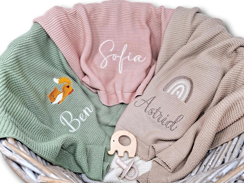 Drei gestrickte Babydecken mit Name und Motiv bestickt. Eine ist lindgrün mit einem Fuchs und dem Namen "Ben", eine ist rosa mit "Sofia" und die letzte ist beige mit einem Regenbogen und "Astrid".