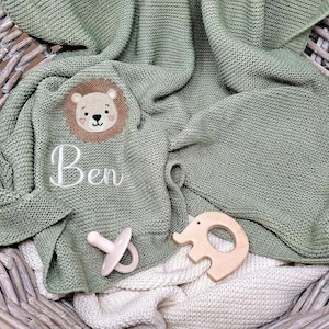 May include: A sage green knitted blanket featuring an embroidered lion face and the name Ben in white script. A pale pink pacifier and a light-colored wooden elephant-shaped teether are placed on top of the textured fabric surface.