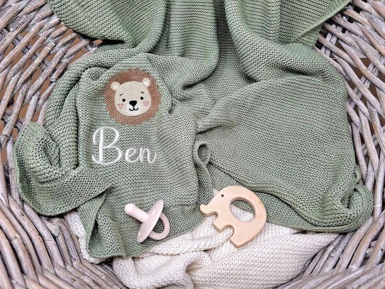 May include: A sage green knitted blanket featuring an embroidered lion face and the name Ben in white script. A pale pink pacifier and a light-colored wooden elephant-shaped teether are placed on top of the textured fabric surface.