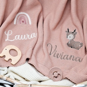 Zwei rosa gestrickte Babydecken mit gestickten Namen und Motiven. Eine Decke hat den Namen "Laura" mit einem Regenbogen, die andere "Viviana" mit einem Reh.